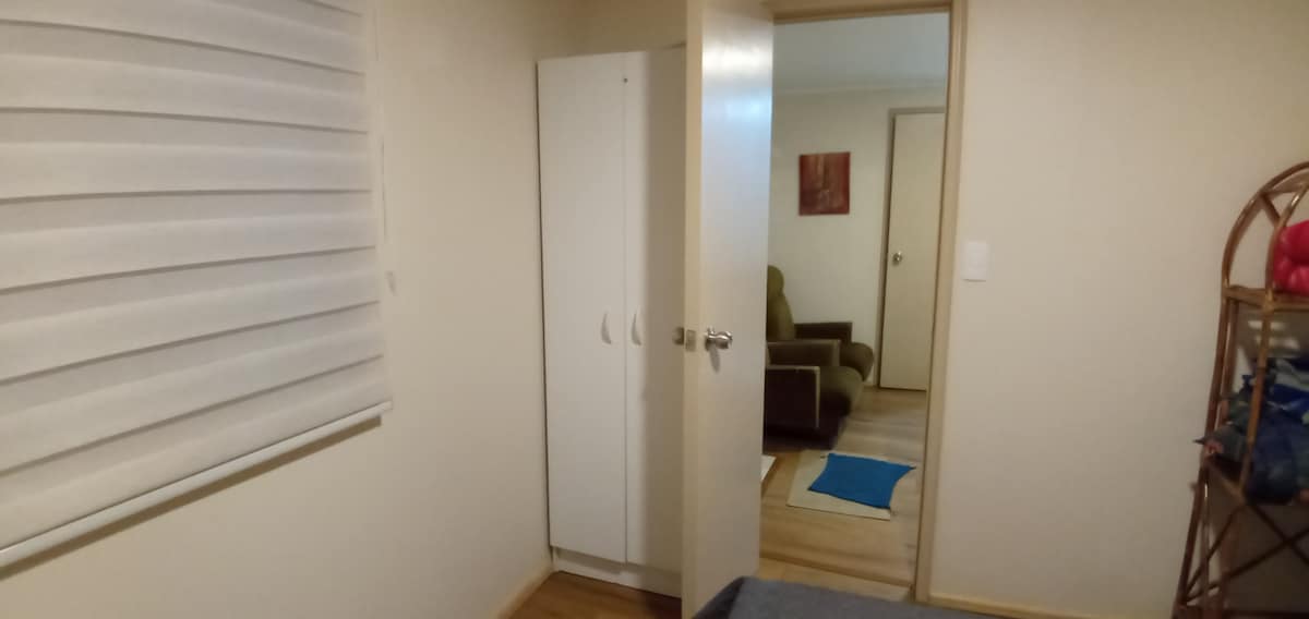 An interior view of the cabin presents a light-colored room with a closed door leading into a living area. A white wardrobe stands against the wall, while a partially visible couch adds to the space's functionality. Natural light filters through a window covered with a light blind.