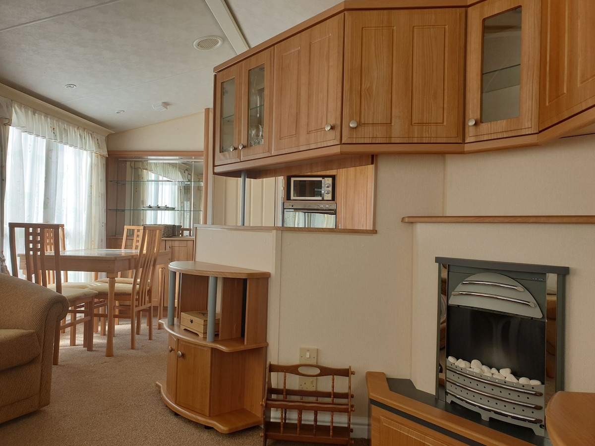 An inviting living area features wooden cabinetry and a functional layout. A cozy seating arrangement includes a light-colored sofa. A dining table is visible nearby, surrounded by wooden chairs. A contemporary fireplace complements the space, with large windows allowing for natural light.