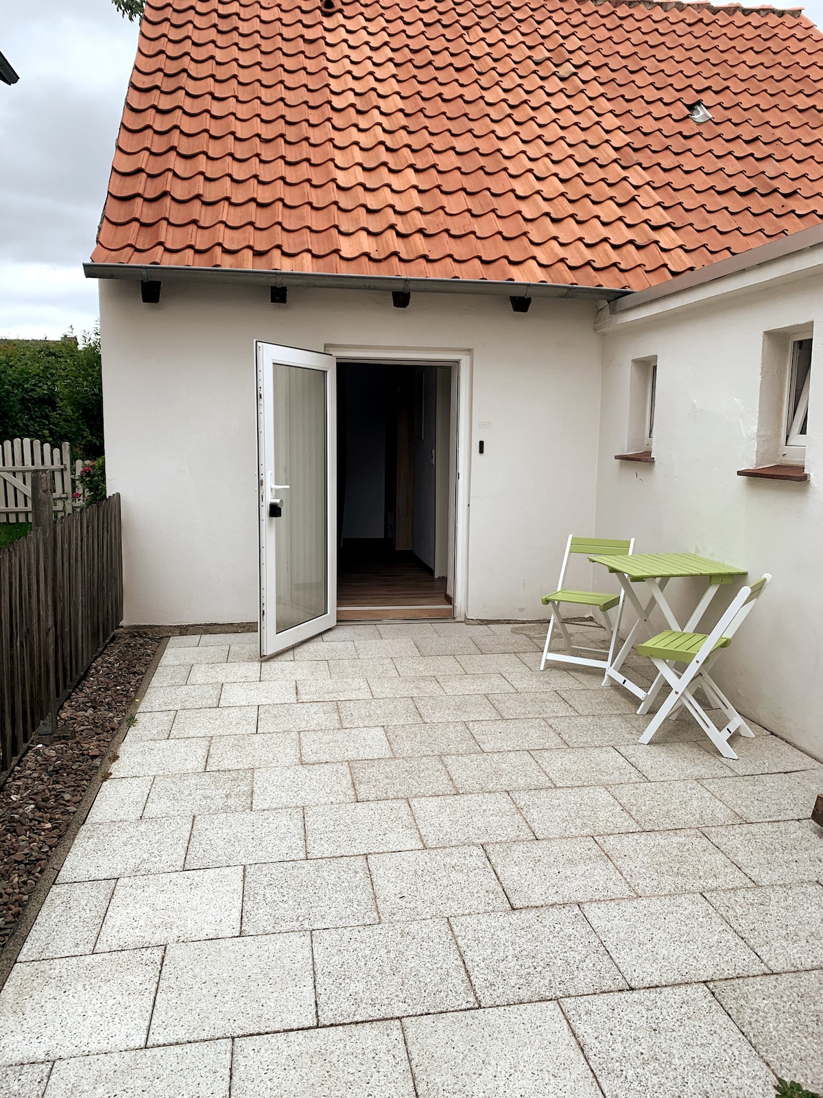 A private outdoor seating area is shown, featuring a small table with two folding chairs. The entrance to the accommodation is visible, framed by a wall with light-colored tiles and a roof with orange shingles. Grass and a fence can be seen in the background.