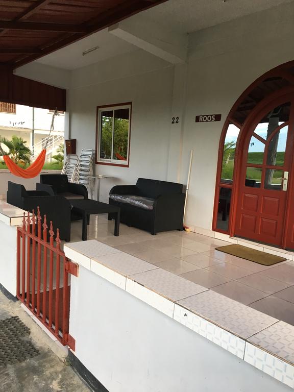An outdoor seating area is visible, featuring two black couches and a matching coffee table on a tiled porch. A red door with a visible number plaque leads into the interior, and a mirror reflects the surrounding greenery. A hammock is also seen in the background.