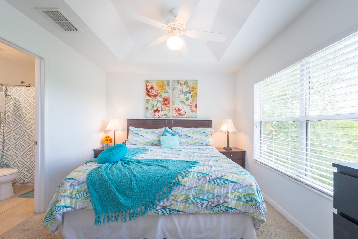 A bright bedroom features a spacious bed adorned with colorful bedding and a throw pillow. Two bedside lamps are positioned on dark nightstands. Natural light flows through large windows, and an ensuite bathroom is visible in the background.