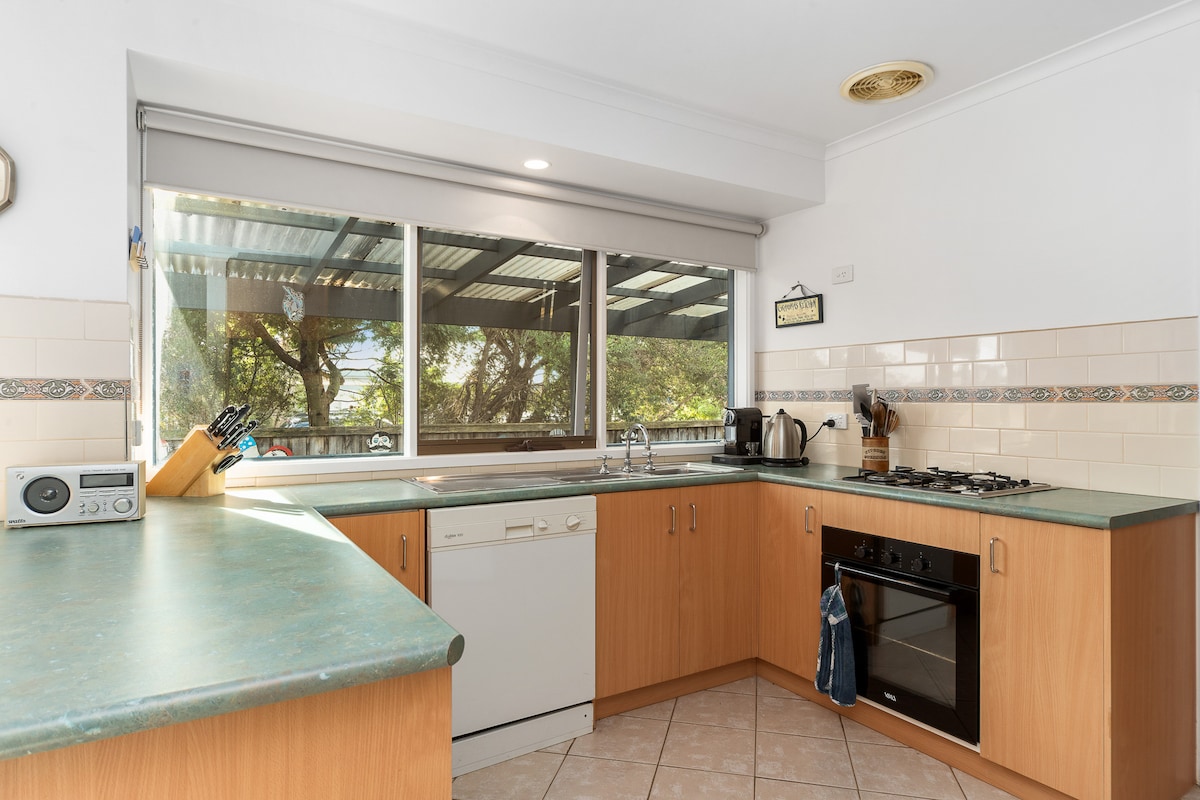 The kitchen features wooden cabinetry and a spacious countertop. Appliances include a dishwasher, oven, and stovetop. Ample natural light is provided by a large window overlooking a shaded outdoor area, creating a functional and well-lit workspace for meal preparation.