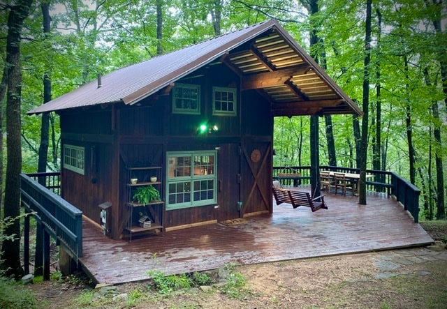 A quaint cabin nestled among trees showcases a wooden exterior with a sloped roof. A spacious deck extends around the cabin, featuring a swing and an outdoor seating area. Large windows allow natural light to brighten the interior, creating a serene and inviting space.