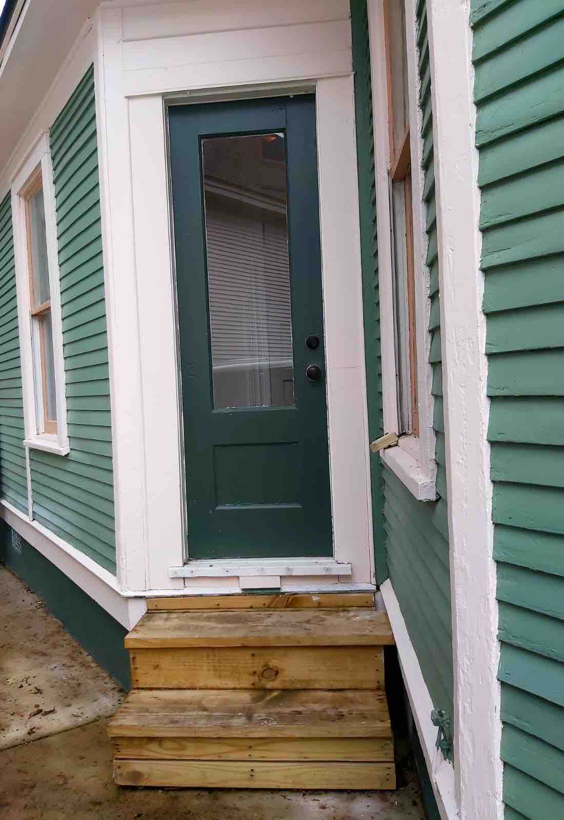 A green exterior wall features a wooden staircase leading up to a door with a large window pane. The door is framed by white trim, providing a contrast to the green siding. Natural light is visible through the door's window.