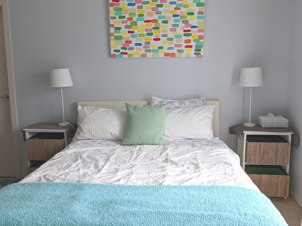 A cozy bedroom features a spacious bed with white linens and a soft turquoise blanket at the foot. Colorful artwork adorns the wall above, while matching bedside tables with lamps sit on either side. Natural light enters through the window, illuminating the room.