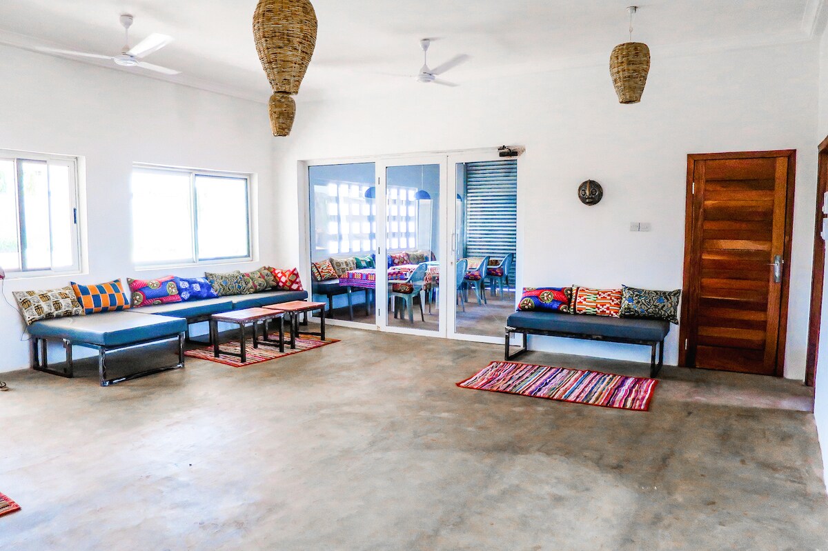 A spacious living area features large windows allowing natural light, with two seating areas consisting of dark sofas adorned with colorful cushions. A striped rug adds texture to the smooth floor, and a wooden door leads to an outside space.