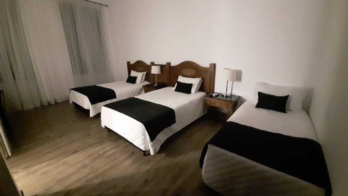 A spacious room is presented with three beds, each covered in crisp white linens with contrasting dark accents. Nightstands with lamps are positioned beside each bed. Expansive windows allow natural light to filter through sheer curtains, providing a bright and airy ambiance.