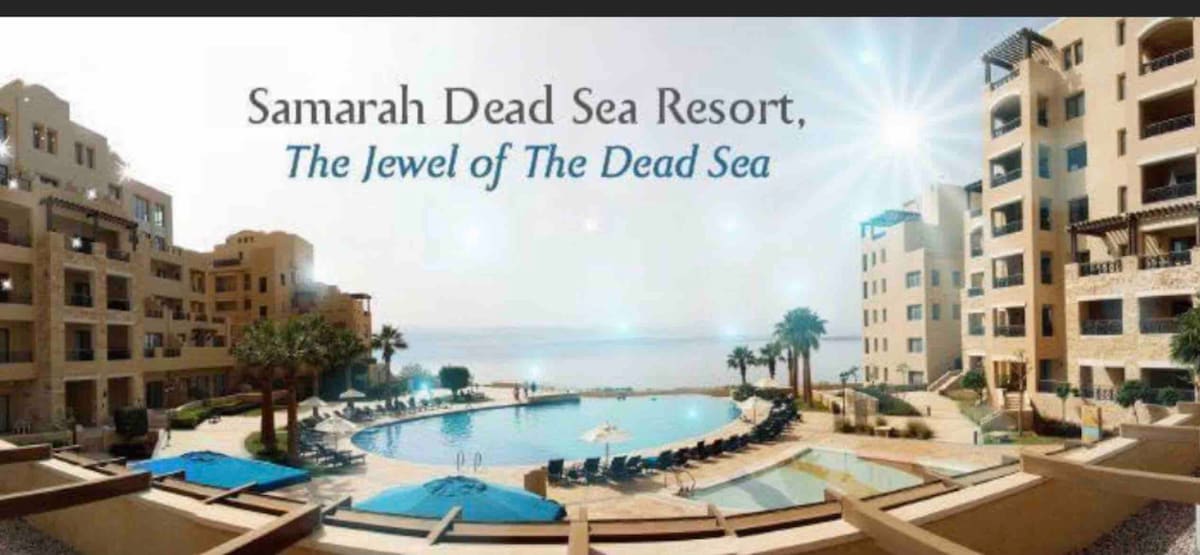 The image displays the Samarah Dead Sea Resort, featuring a large outdoor pool surrounded by lounge chairs and palm trees. The serene sea is visible in the background, with buildings designed in a traditional style framing the scene. Clear skies enhance the overall view.