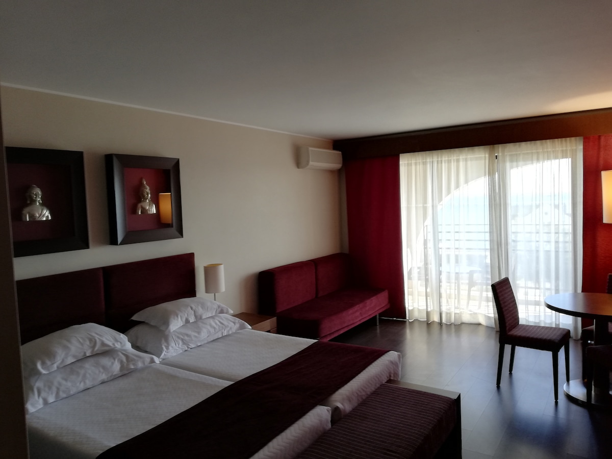 A spacious room features two large beds with soft linens, complemented by a maroon color scheme. Natural light enters through large glass doors that open to a balcony. A comfortable seating area includes a red couch and a small dining table with a chair.