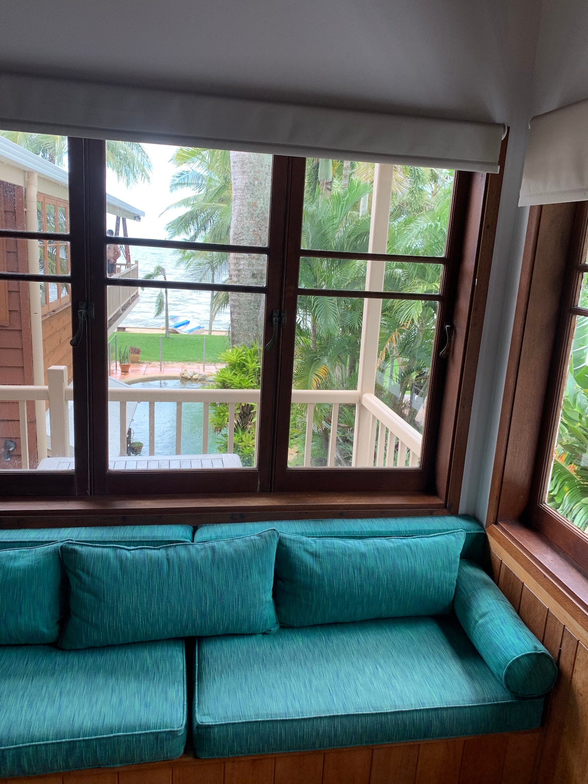 A cozy seating area features a turquoise couch set against large wooden windows. The view includes a tropical landscape with palm trees and a glimpse of the pool area below, offering a serene and tranquil atmosphere.