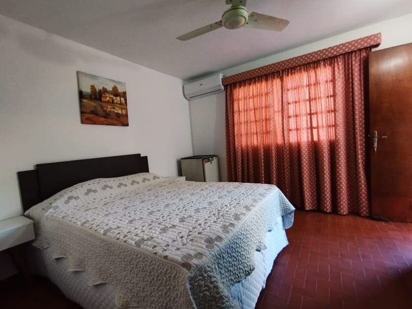 A spacious sleeping area features a large bed covered with a light-colored quilt. A ceiling fan adds to the comfort, while a small refrigerator is positioned nearby. Natural light filters through the patterned curtains, creating a cozy ambiance.