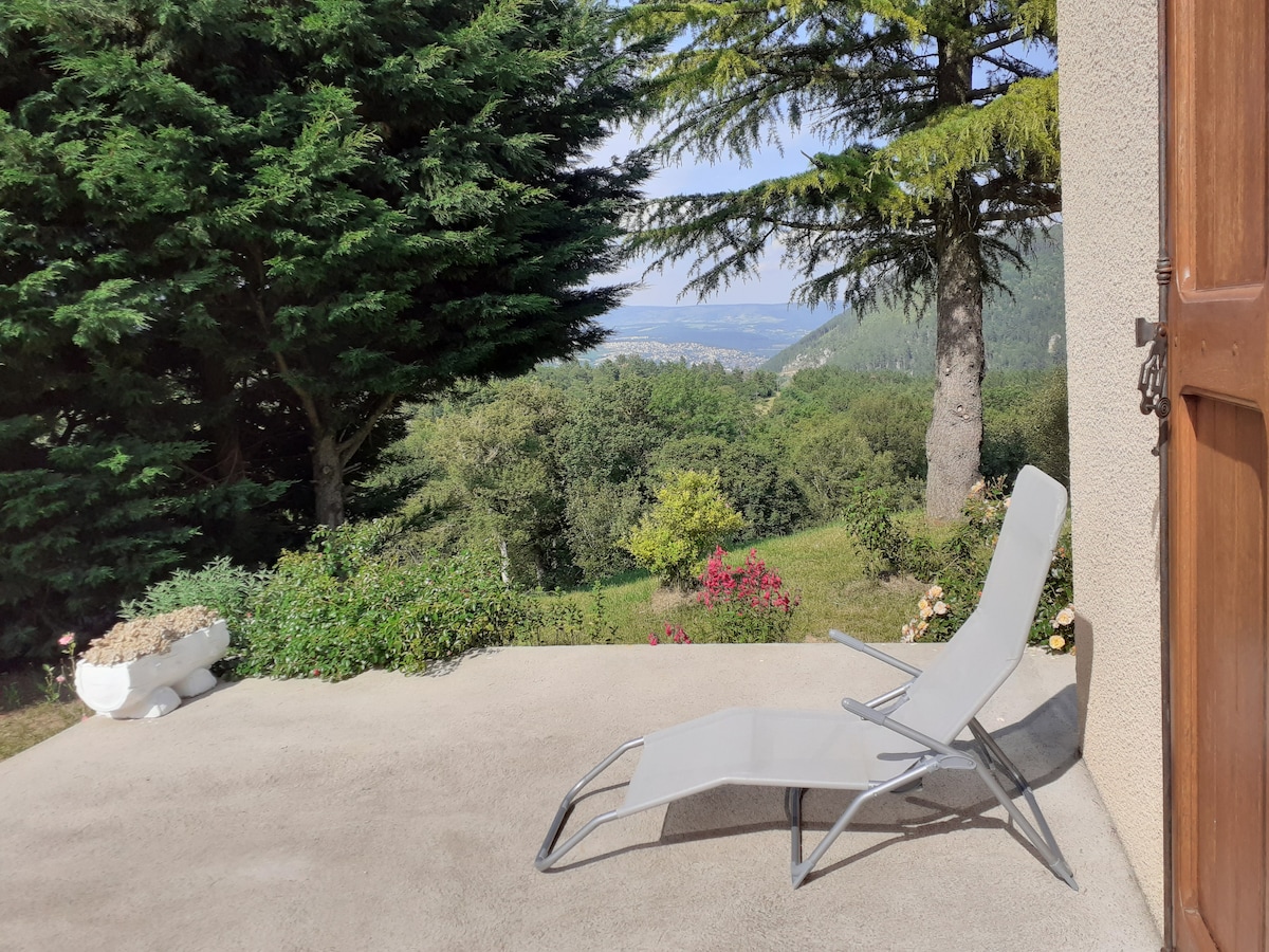 A sunlit terrace is shown, featuring a light gray lounge chair positioned for relaxation. Surrounded by green trees and blooming flowers, the view extends over the landscape, showcasing a serene countryside and distant hills.