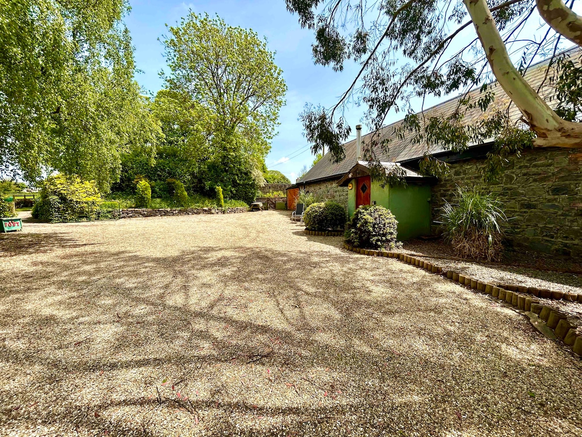A spacious gravel area is shown, bordered by lush greenery and trees. A charming barn structure is nestled nearby, complemented by well-maintained garden areas. Soft shadows are cast by the trees, creating a serene and open outdoor environment.