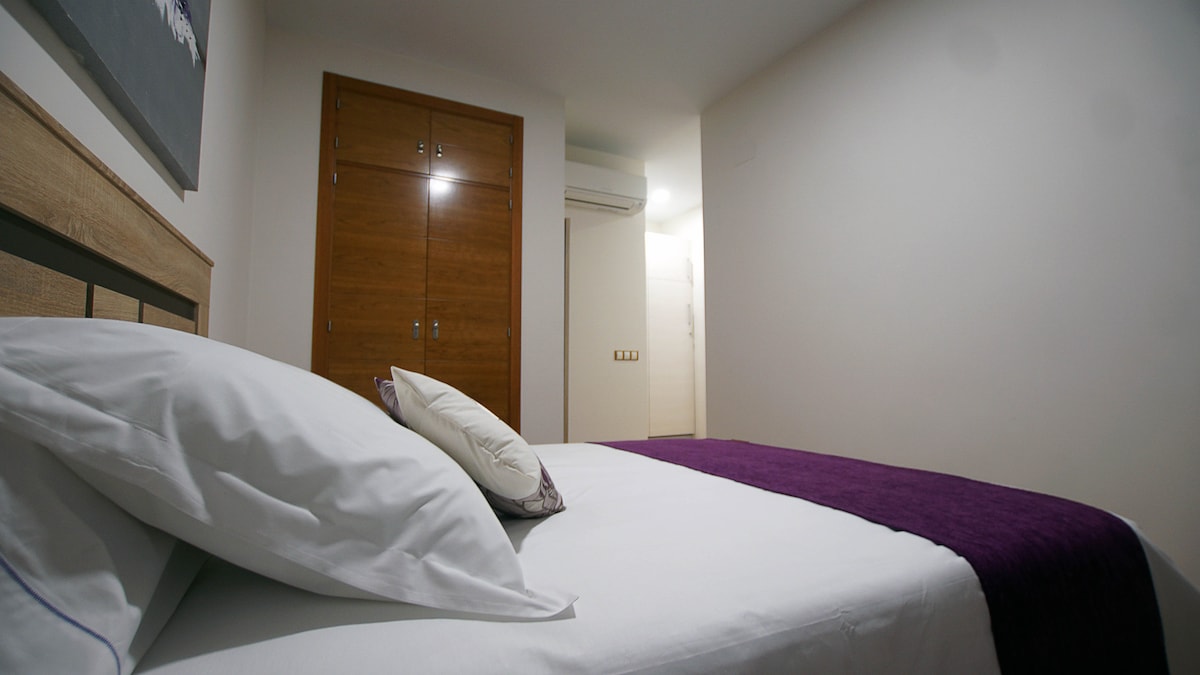 A well-lit bedroom features a spacious bed with a white duvet and a purple throw blanket. A decorative pillow rests against the headboard. Wooden wardrobes are visible in the background, while a door leads to another area of the apartment.