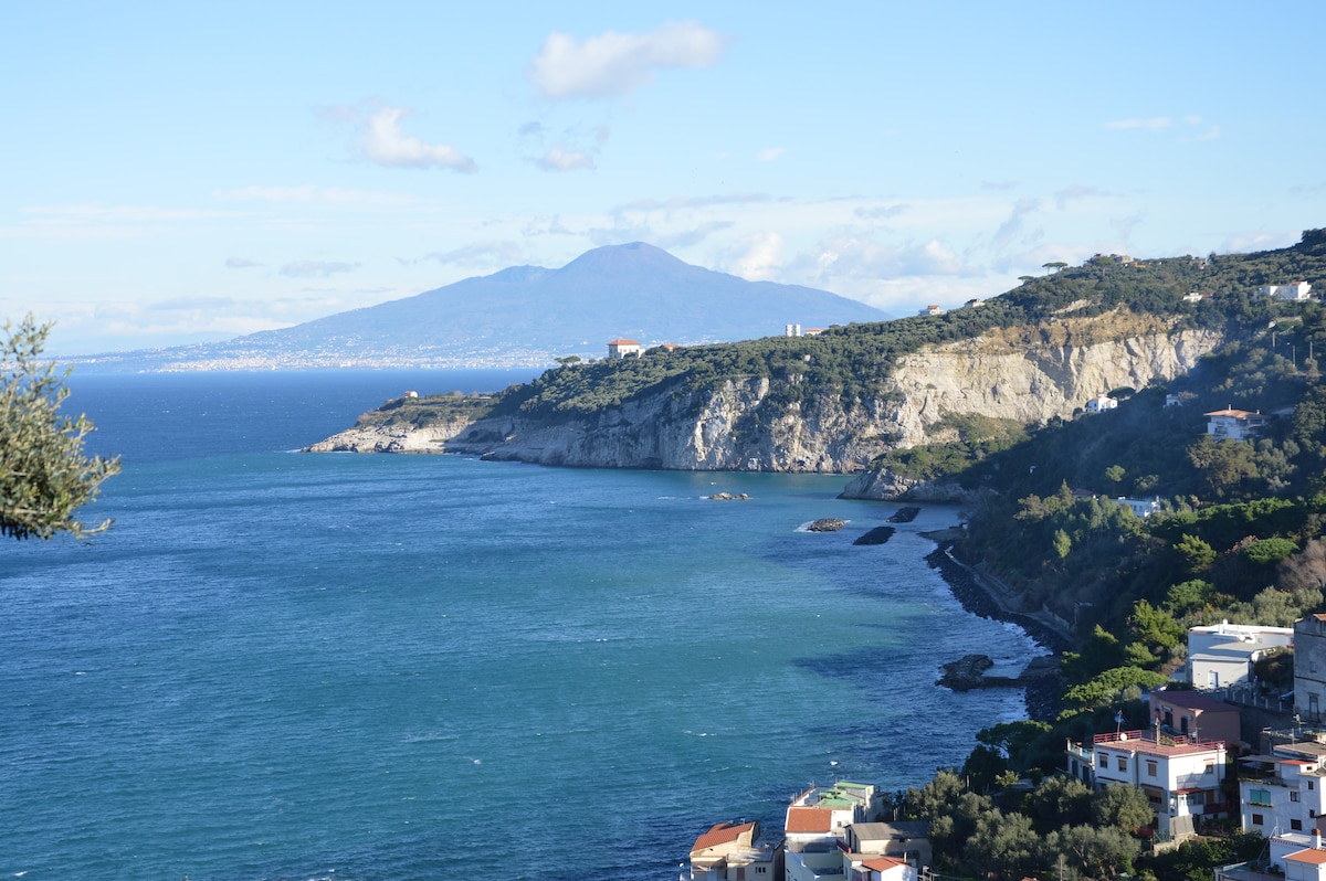 A breathtaking view of the Gulf of Naples is presented, with rugged cliffs and verdant hills extending along the coastline. The vibrant blue waters contrast with the earthy tones of the land. In the distance, the silhouette of a mountain adds depth to the picturesque landscape.