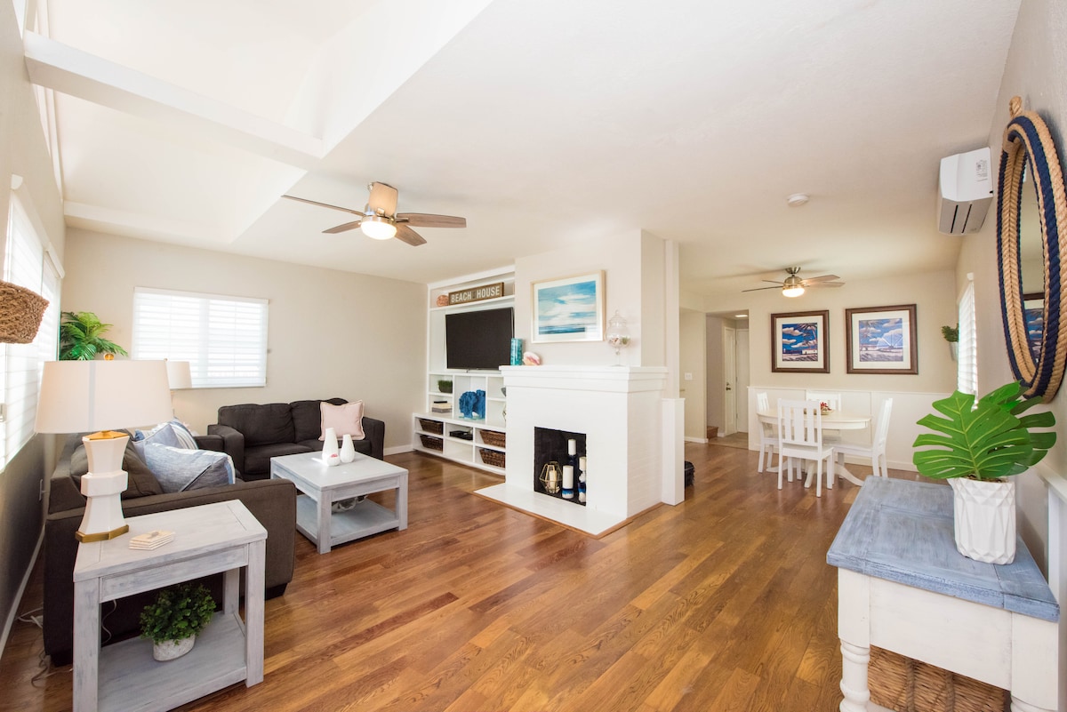 The living room is characterized by hardwood flooring and features a dark sofa and a light coffee table. A TV is mounted above a modern fireplace. A dining area is visible with a white table and chairs. Natural light enters through windows with blinds.