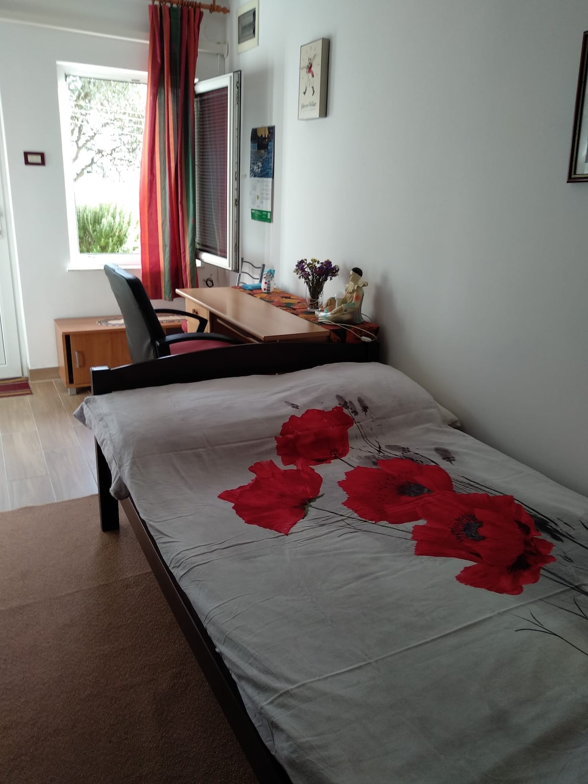 A cozy sleeping area features a single bed adorned with a floral-patterned blanket in shades of white and red. A desk and chair are positioned near a window, allowing natural light to illuminate the space. Red curtains frame the window, adding a touch of color.