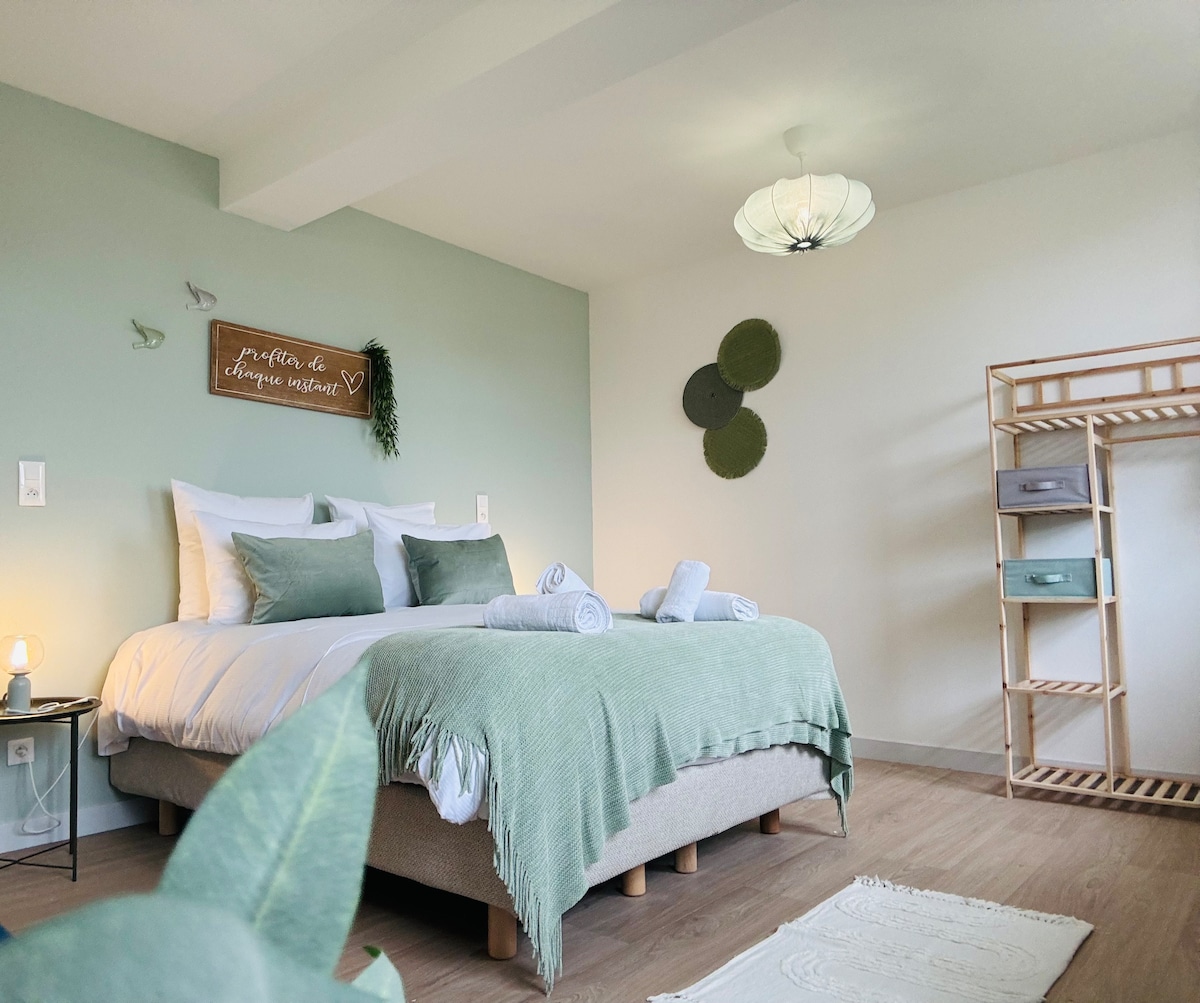 A serene bedroom features a spacious bed dressed in soft green linens. Decorative accents include a wall sign and wall art, complemented by a minimalist wooden shelving unit. Natural light fills the room, highlighting the calming color palette for a restful ambiance.