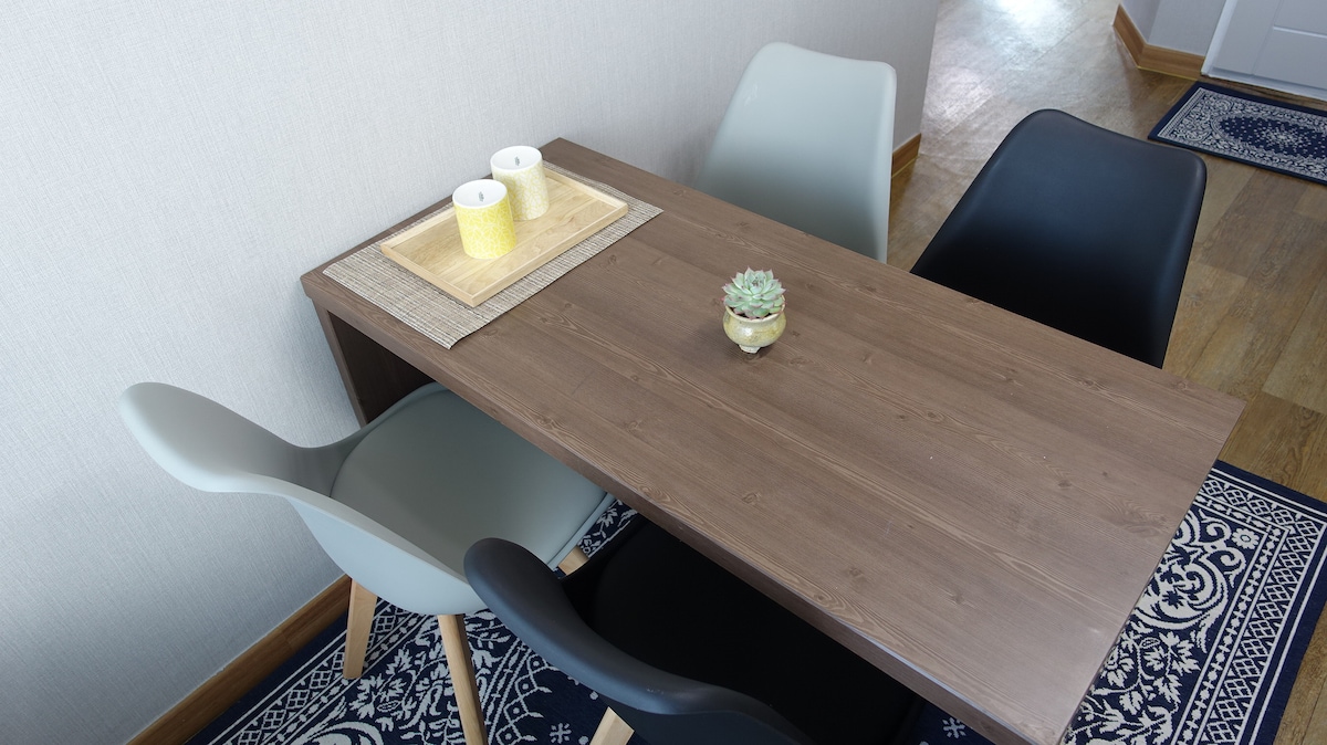 A modern dining table is displayed, featuring a smooth surface with a decorative centerpiece. Two light-colored chairs and two dark chairs are arranged around the table. Natural light brightens the area, with a textured rug visible beneath.