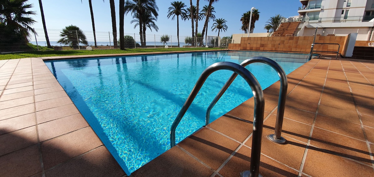 A tranquil swimming pool features crystal-clear water, with steps leading down into it. Surrounding palm trees sway gently, creating a serene environment. The pool offers direct views of the beach and sea, enhancing the outdoor experience.