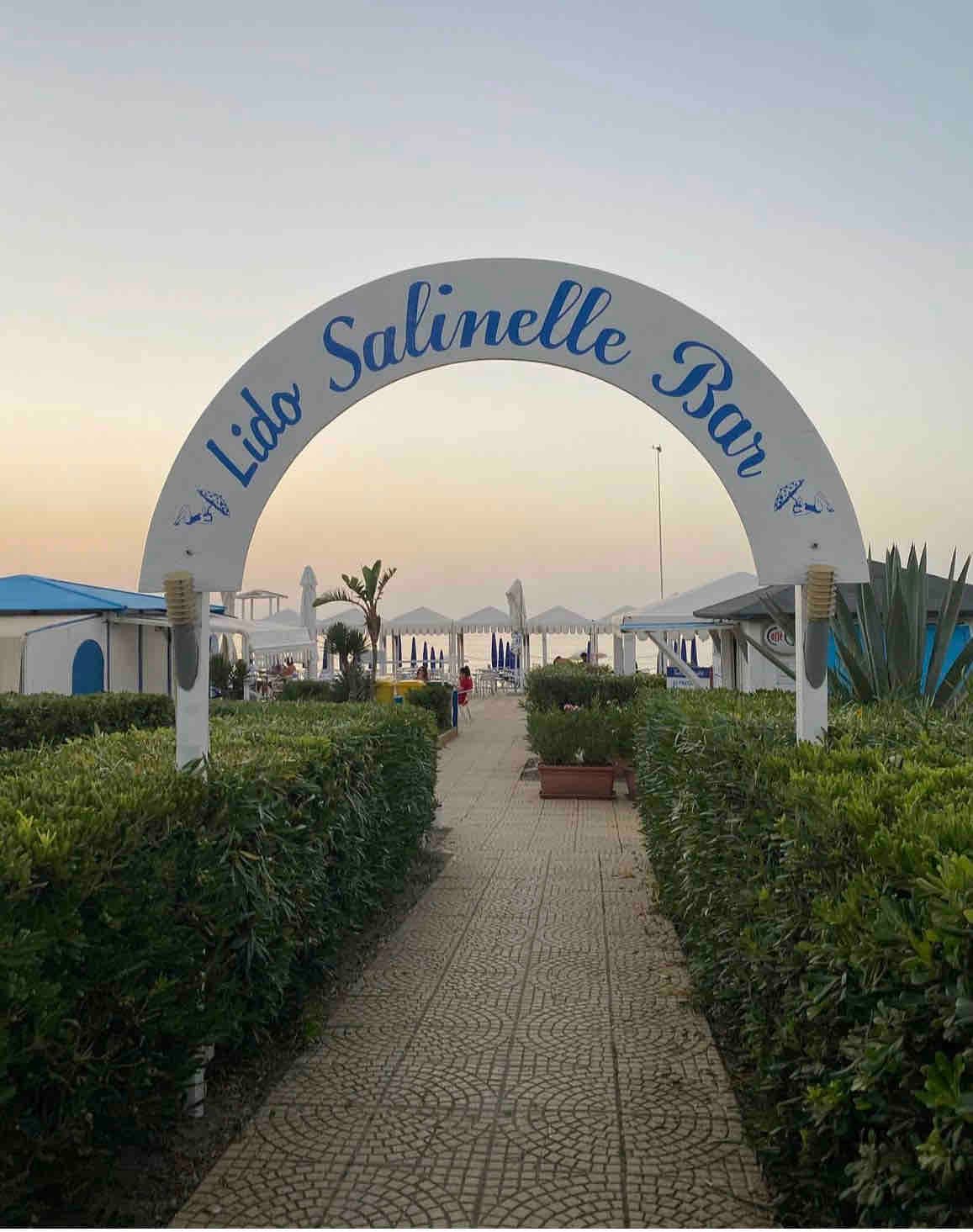 An archway marked 'Lido Salinelle Bar' welcomes visitors, leading through well-maintained greenery towards the beach. The path is paved with stone, guiding towards sun loungers and umbrellas, with the sea visible in the distance, framed by a soft twilight sky.