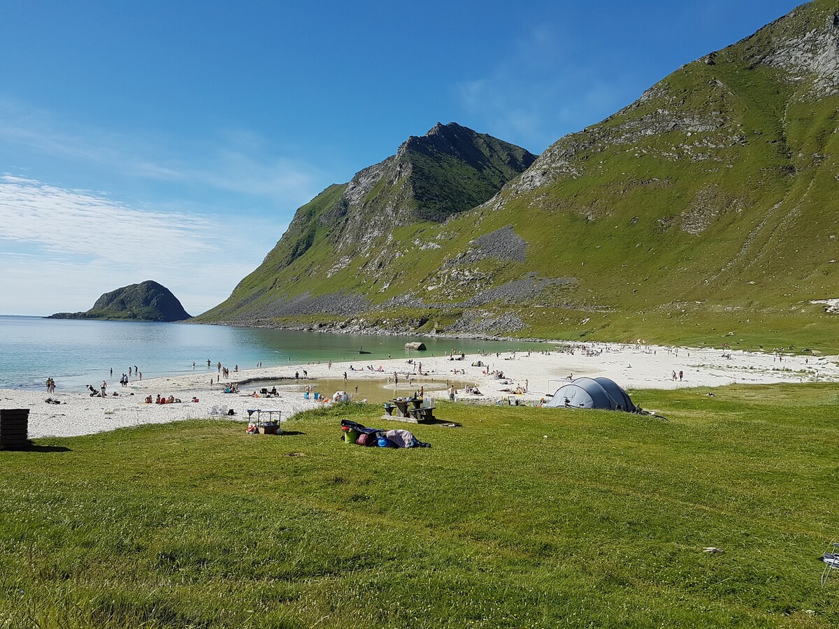 A serene beach scene features soft white sand and clear blue waters, framed by lush green hills. Groups of people can be seen enjoying the beach, while colorful towels and blankets are spread across the grassy area. Gentle waves lap at the shore under a clear sky.