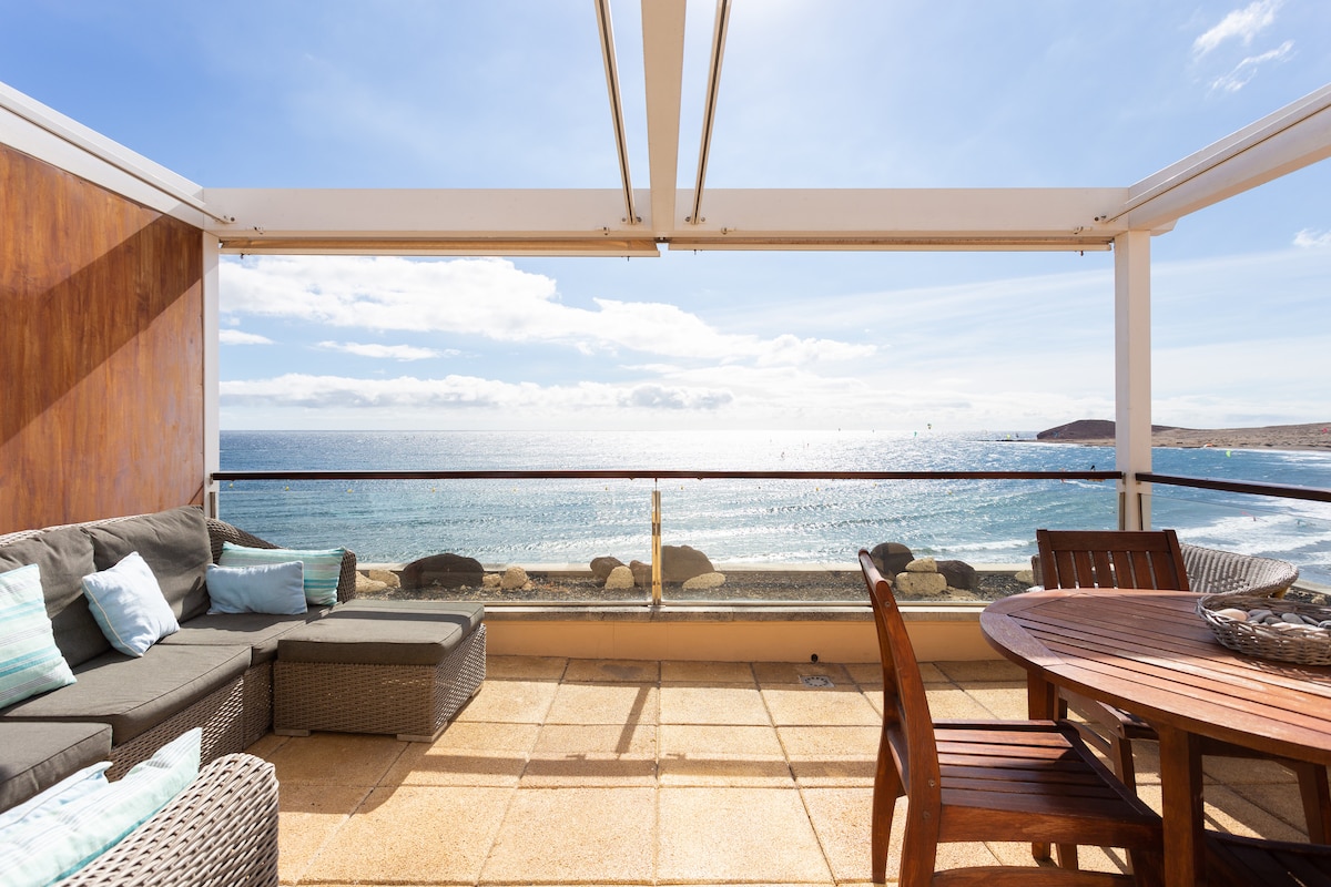 A spacious terrace offers stunning ocean views, featuring a comfortable seating area with soft cushions and a wooden dining table. The outdoor space is bathed in natural light, with the sea visible in the distance and the sky showcasing a clear blue hue.