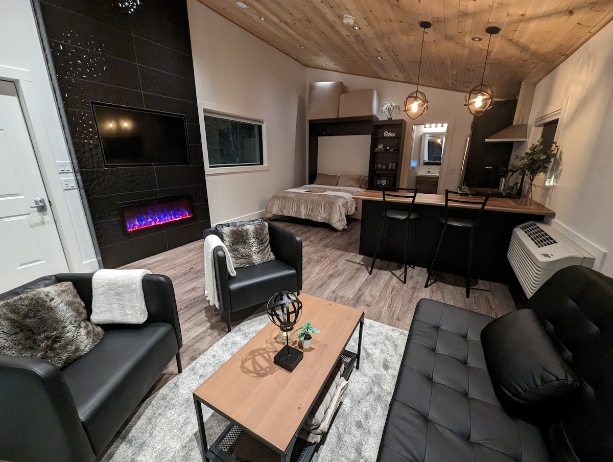 A modern interior featuring a cozy sitting area with two black chairs and a coffee table. A sleek electric fireplace is mounted on the wall, while a sleeping area with a spacious bed is visible in the background. A kitchen counter with bar stools complements the space.
