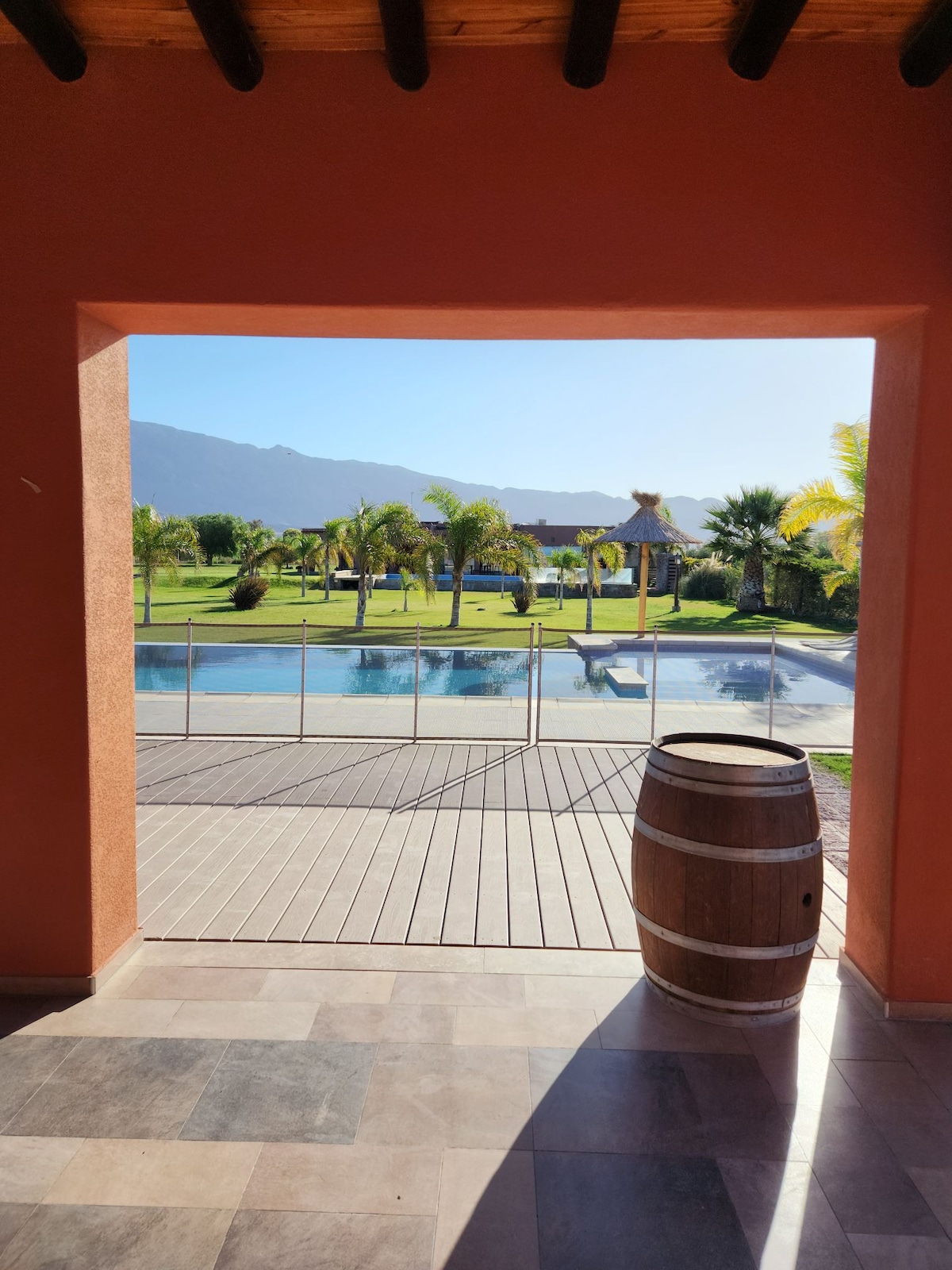 A view from an archway showcases a large private yard featuring a glistening pool and tropical palm trees. A wooden barrel is positioned in the foreground, while the backdrop displays lush greenery and distant mountains under a clear sky.