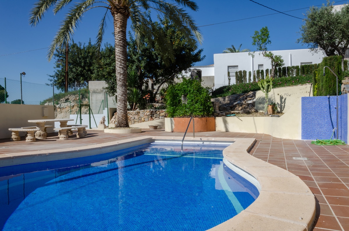A tranquil swimming pool is framed by palm trees and surrounding greenery. The pool reflects a clear blue sky, with a tiled deck providing ample space for seating. Nearby, a rustic stone table and benches are positioned for relaxation, contributing to the serene outdoor environment.