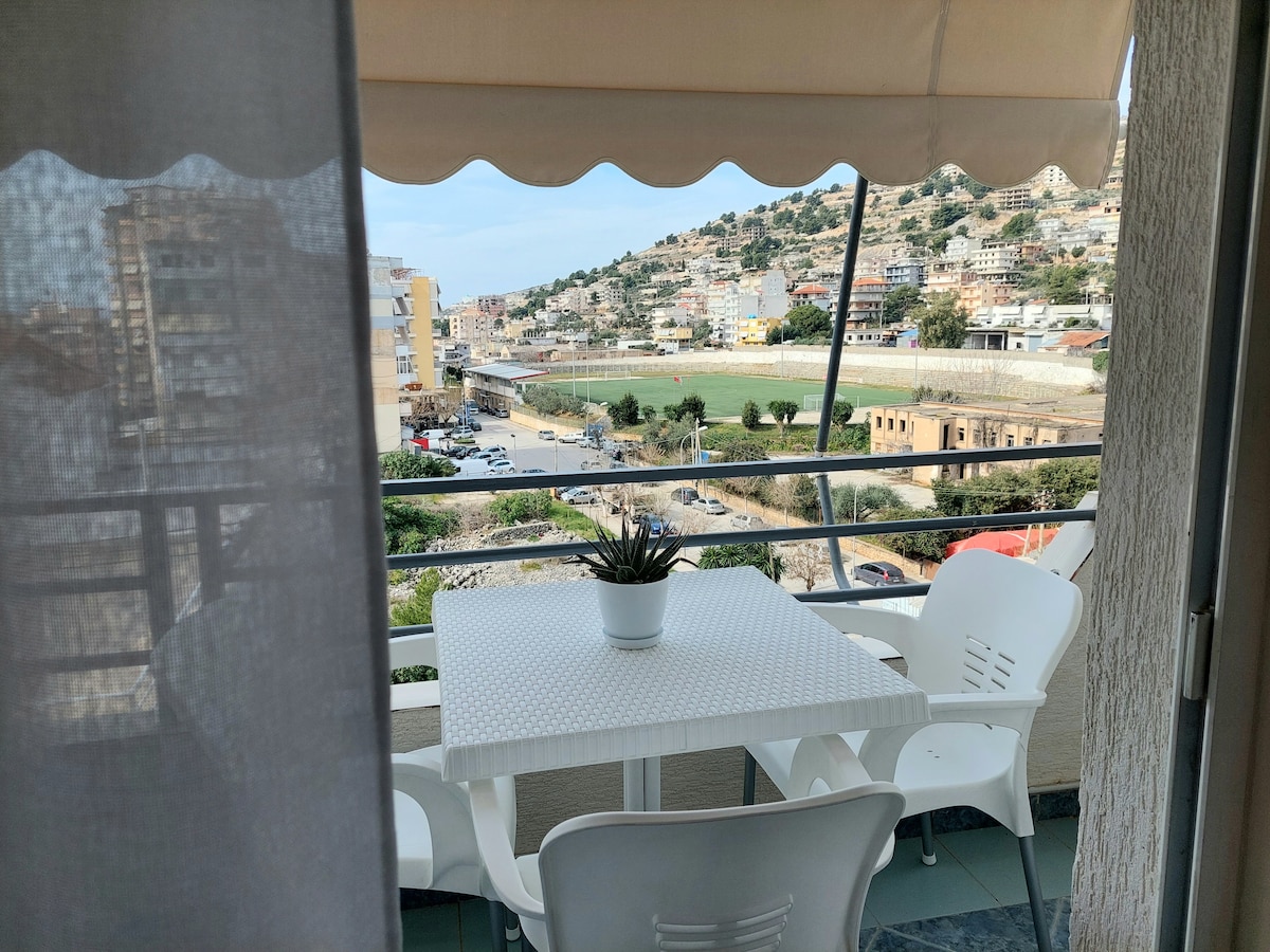 A spacious balcony features a white table and chairs, providing a comfortable spot to enjoy meals or relax. Scenic views of the surrounding hills and cityscape are visible, framed by a soft awning. A small potted plant adds a touch of greenery to the setting.