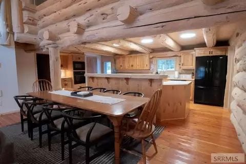 A spacious dining area features a long wooden table surrounded by black chairs, with warm wood cabinetry in the background. An island counter stands in the kitchen, complemented by modern appliances. Natural light enters through a nearby window, enhancing the rustic wooden beams overhead.