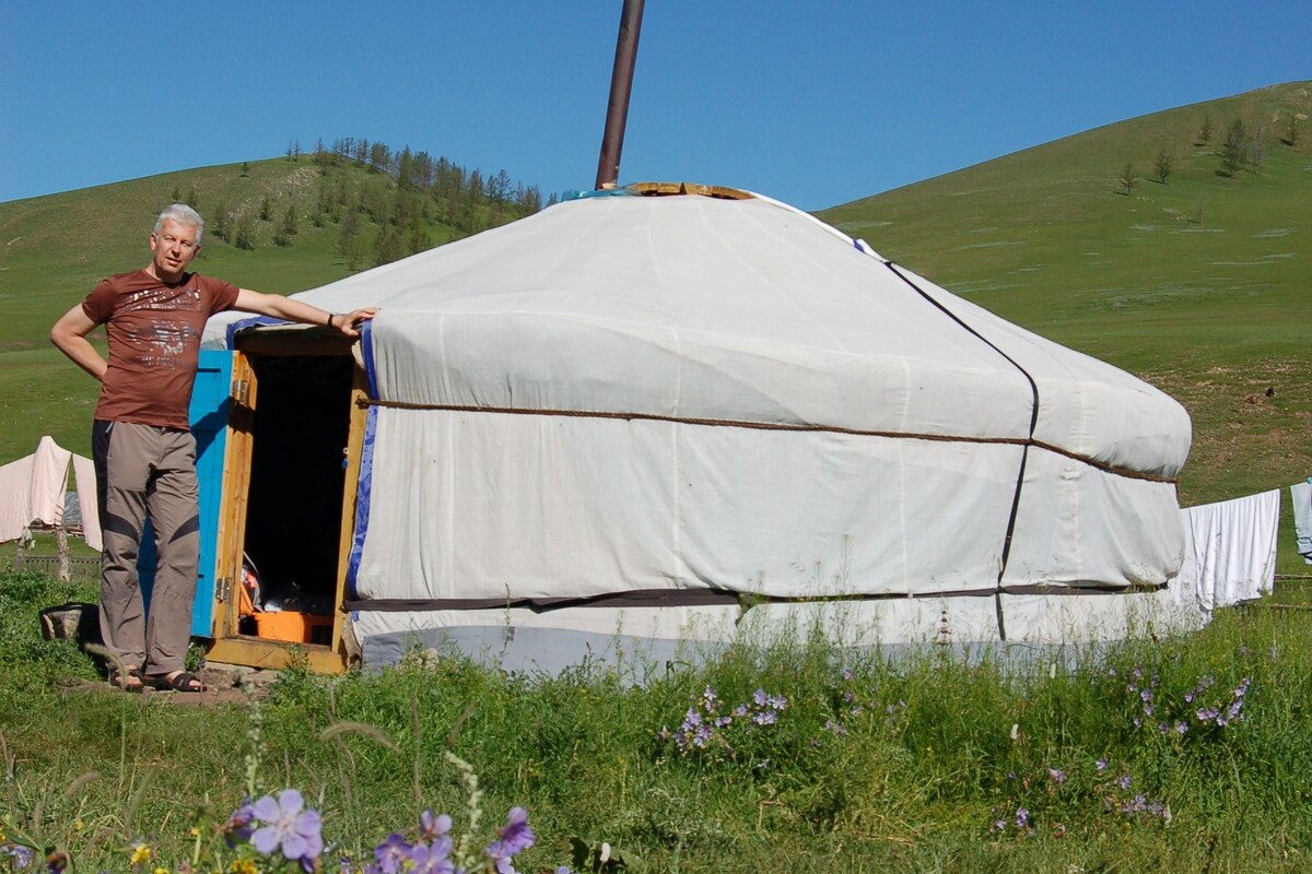 A traditional ger, covered in white felt, stands on a grassy landscape with gentle green hills in the background. The entrance is framed with blue, highlighting the simplicity of nomadic living. Nearby, clotheslines with items are visible, adding to the rustic charm of the setting.