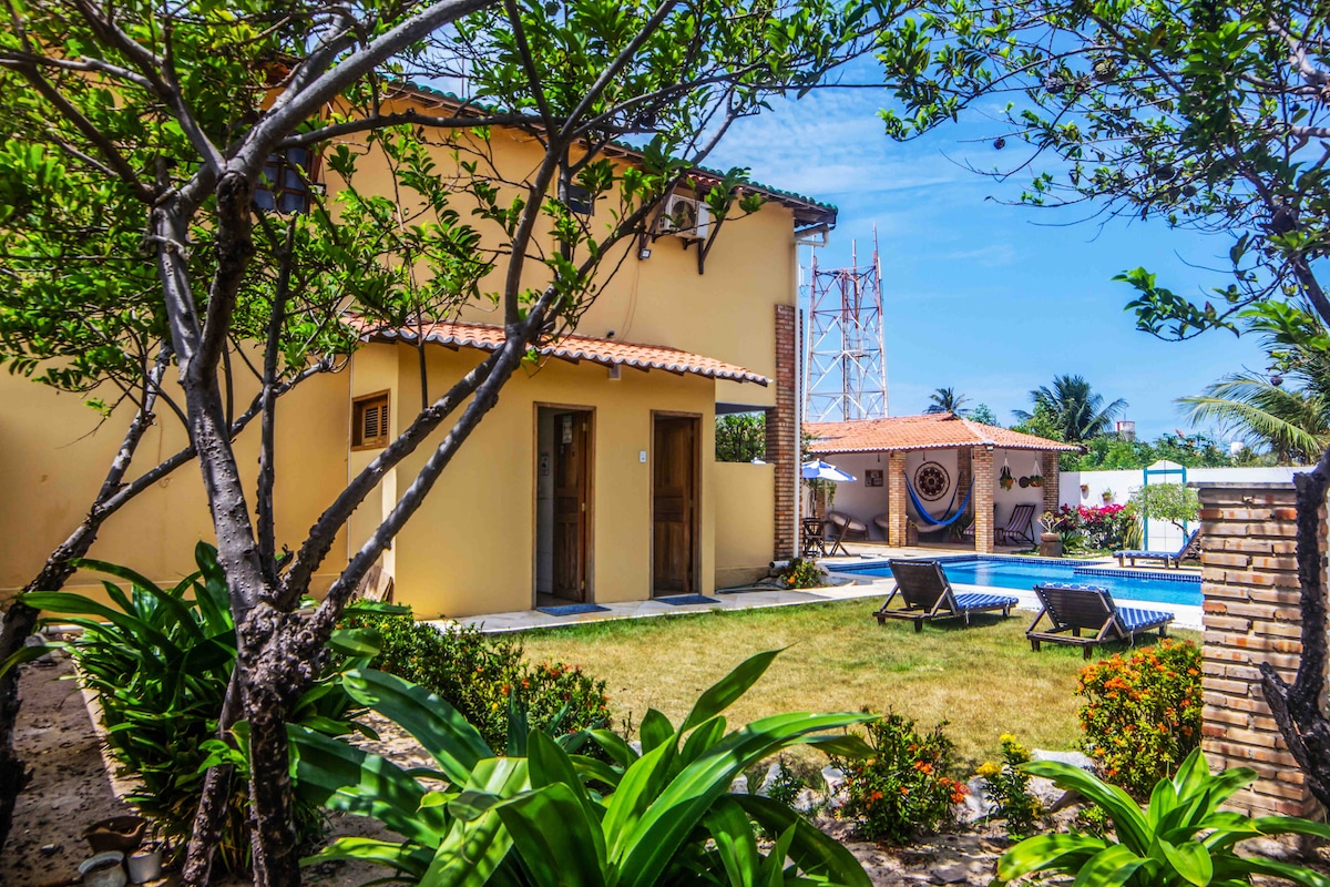 A well-maintained garden surrounds a charming two-story house. Features include a swimming pool and two sun loungers positioned on the grass. Tropical plants and trees provide shade and greenery, while the house showcases a bright yellow exterior and welcoming entryways.