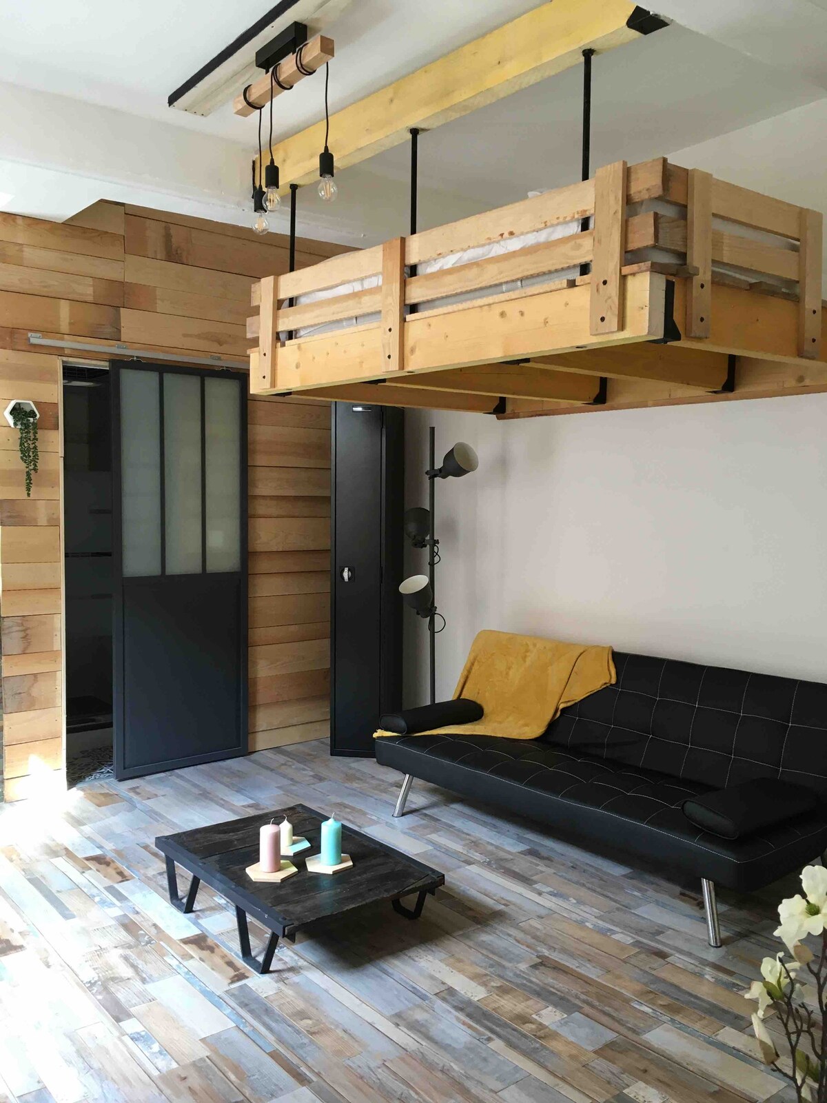 A cozy studio space features a wooden lofted bed suspended above a sleek black sofa. A small coffee table sits in front, showcasing colorful candles. Natural wood paneling creates a warm backdrop, with an industrial-style sliding door providing access to an adjoining room.