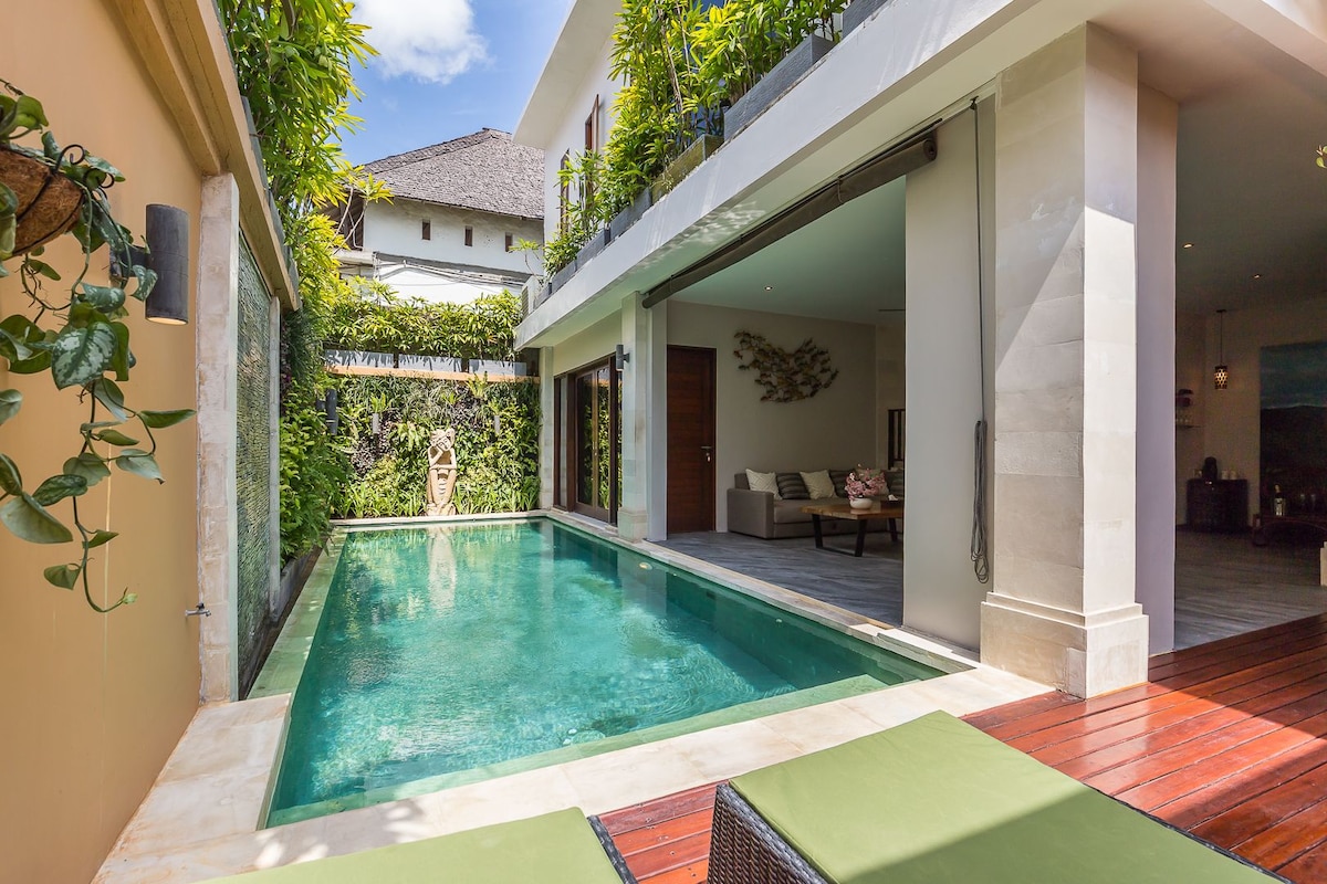 A private swimming pool is framed by lush greenery and natural stone walls. The refreshing water reflects soft sunlight, while comfortable sun loungers are positioned nearby, inviting relaxation. Open spaces connect the pool area to the airy living room, enhancing the seamless indoor-outdoor flow.