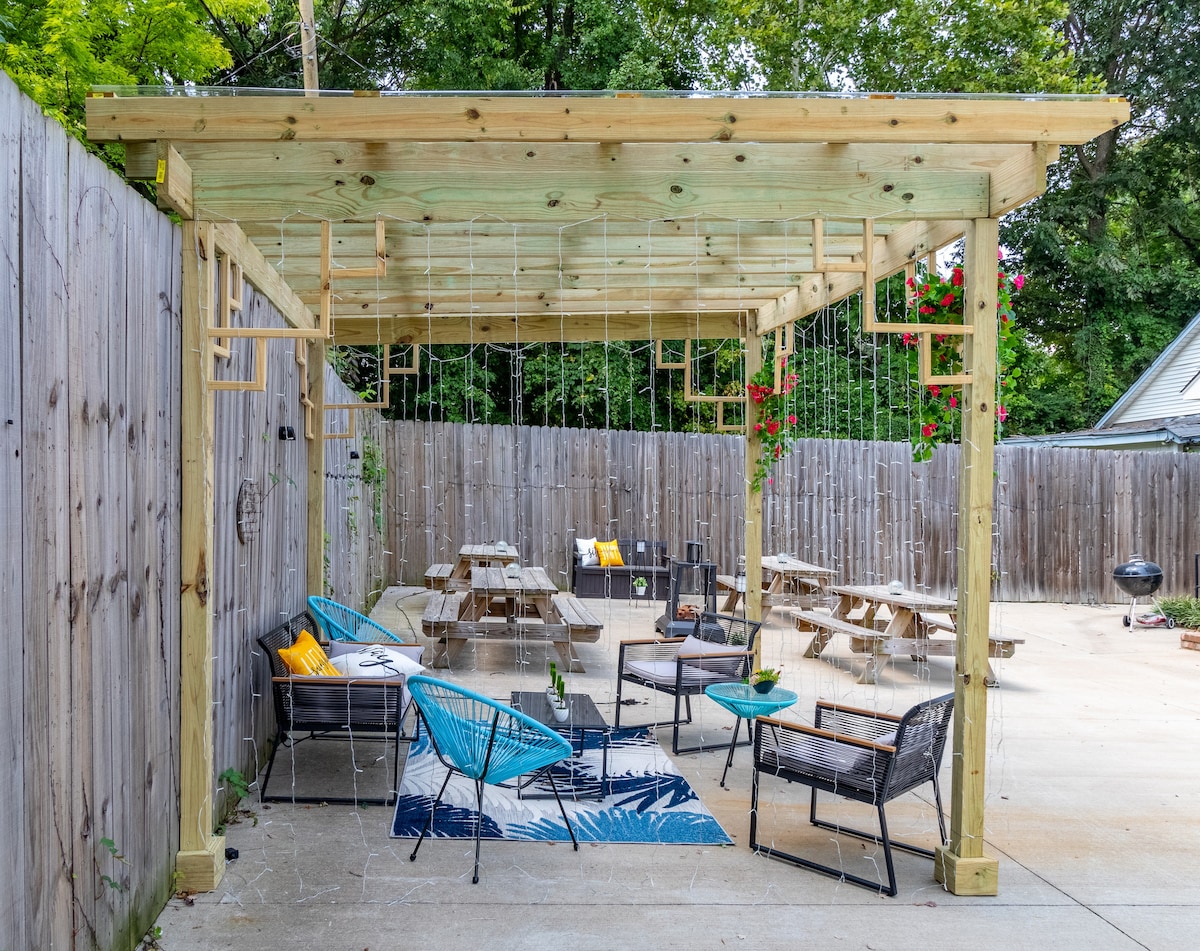 A shared backyard space is highlighted by a wooden pergola providing partial shade. Multiple picnic tables are surrounded by various seating options, including chairs with vibrant cushions. Hanging plants add a touch of greenery, while a decorative rug defines a cozy sitting area.
