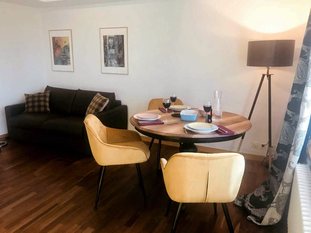 The image features a dining area with a round wooden table set for a meal, including plates and glasses. Yellow chairs surround the table. A dark sofa with plaid cushions is positioned against the wall, and a floor lamp provides soft lighting, enhancing the inviting space.