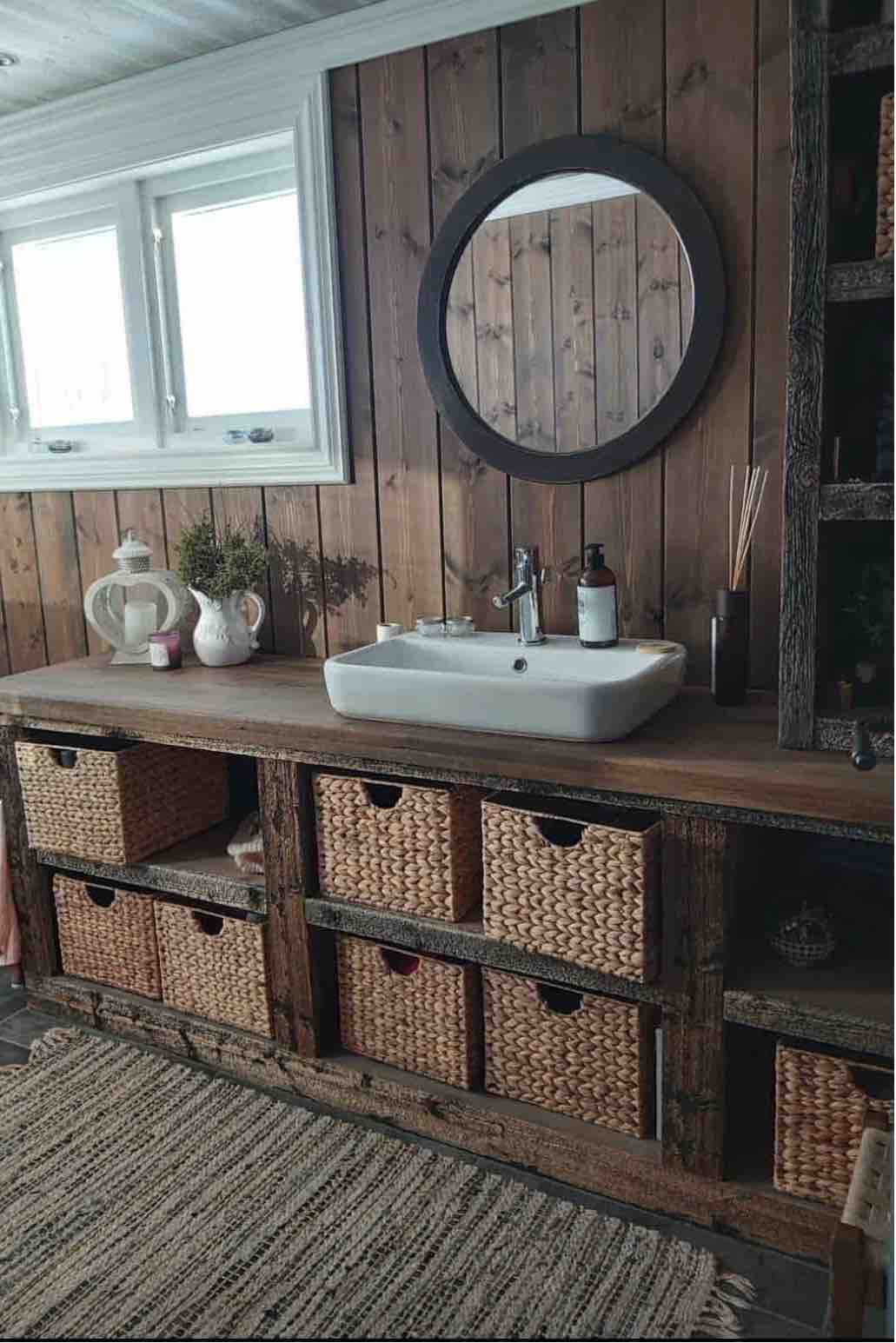 A modern bathroom is showcased with a stylish wooden vanity featuring a white sink. The space is enhanced by a round mirror above the sink and natural light streaming through two windows. Woven baskets are neatly arranged below, providing ample storage.