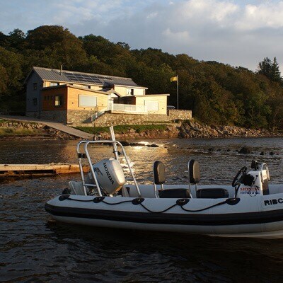 The villa is visible in the background, showcasing a modern design with solar panels on the roof and large windows. A small, motorized boat is parked in the foreground, positioned near a jetty that extends into the water. The surrounding landscape features trees and a tranquil body of water.