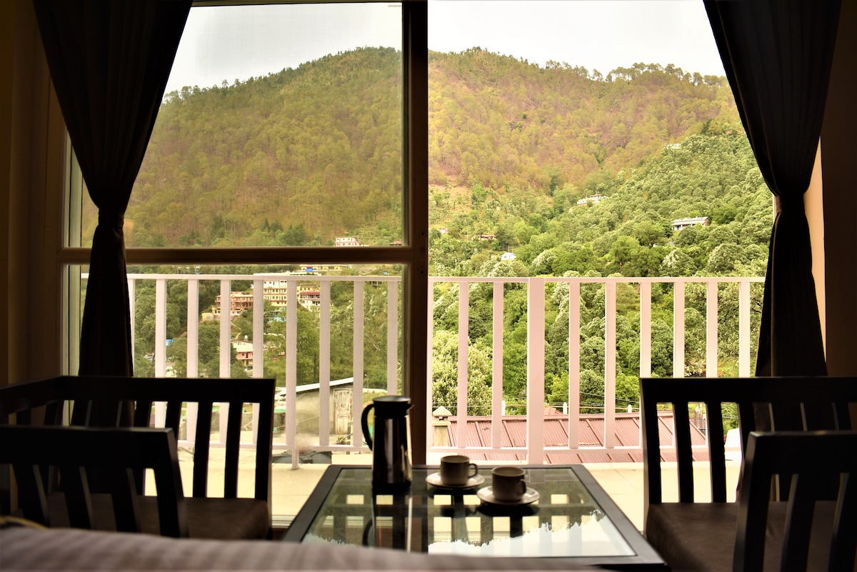 A spacious room is visible through large glass doors, offering an expansive view of lush green hills. Two chairs and a glass table are situated near the window, with a kettle and two cups placed on the table. Natural light filters in, enhancing the serene atmosphere.