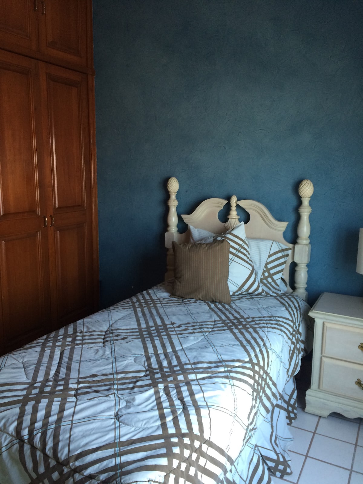A cozy bedroom is presented with a single bed featuring a decorative headboard. The bedding displays a subtle checkered pattern in light and dark tones. A lamp is situated on a nightstand beside the bed, while a wooden wardrobe occupies one wall.