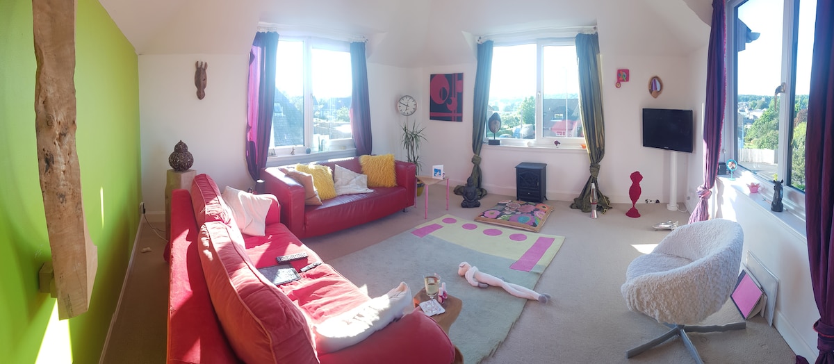 A spacious living area features large windows inviting natural light, offering views of the surrounding landscape. Colorful furniture, including a red sofa and unique armchair, adds character. A playful rug is positioned on soft carpeting, while decorative elements and plants enhance the inviting atmosphere.