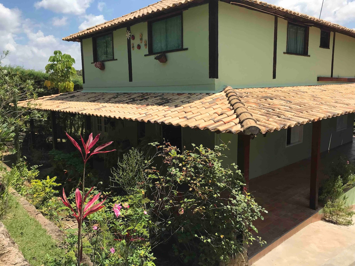 The exterior of a two-story house is characterized by a sloped tiled roof and shaded porch. Lush plants and flowers surround the entrance, enhancing the natural landscape. Large windows allow light to enter the upper floor, providing an inviting view of the surroundings.