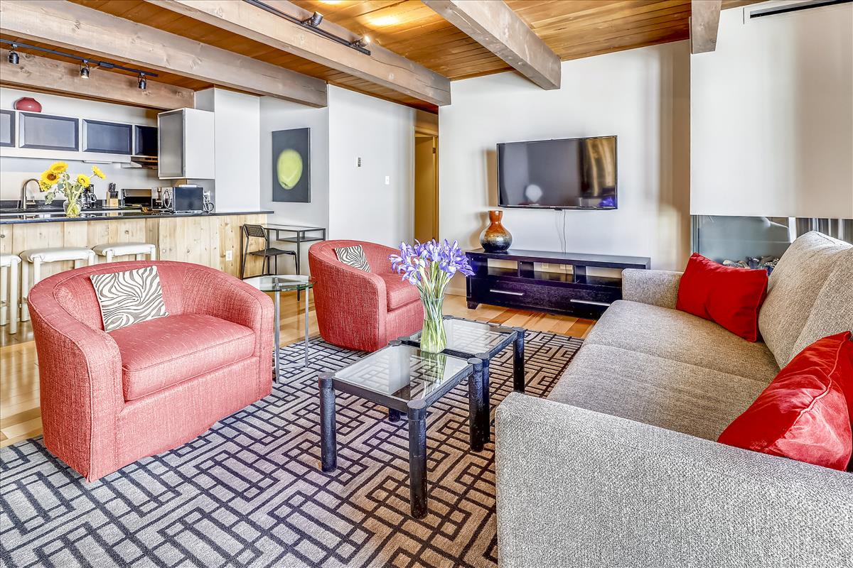 A bright living area features two red accent chairs and a spacious gray sofa arranged around a glass coffee table. A flat-screen TV is mounted on the wall, with a modern kitchen area visible in the background, showcasing a rich wood finish and contemporary appliances.