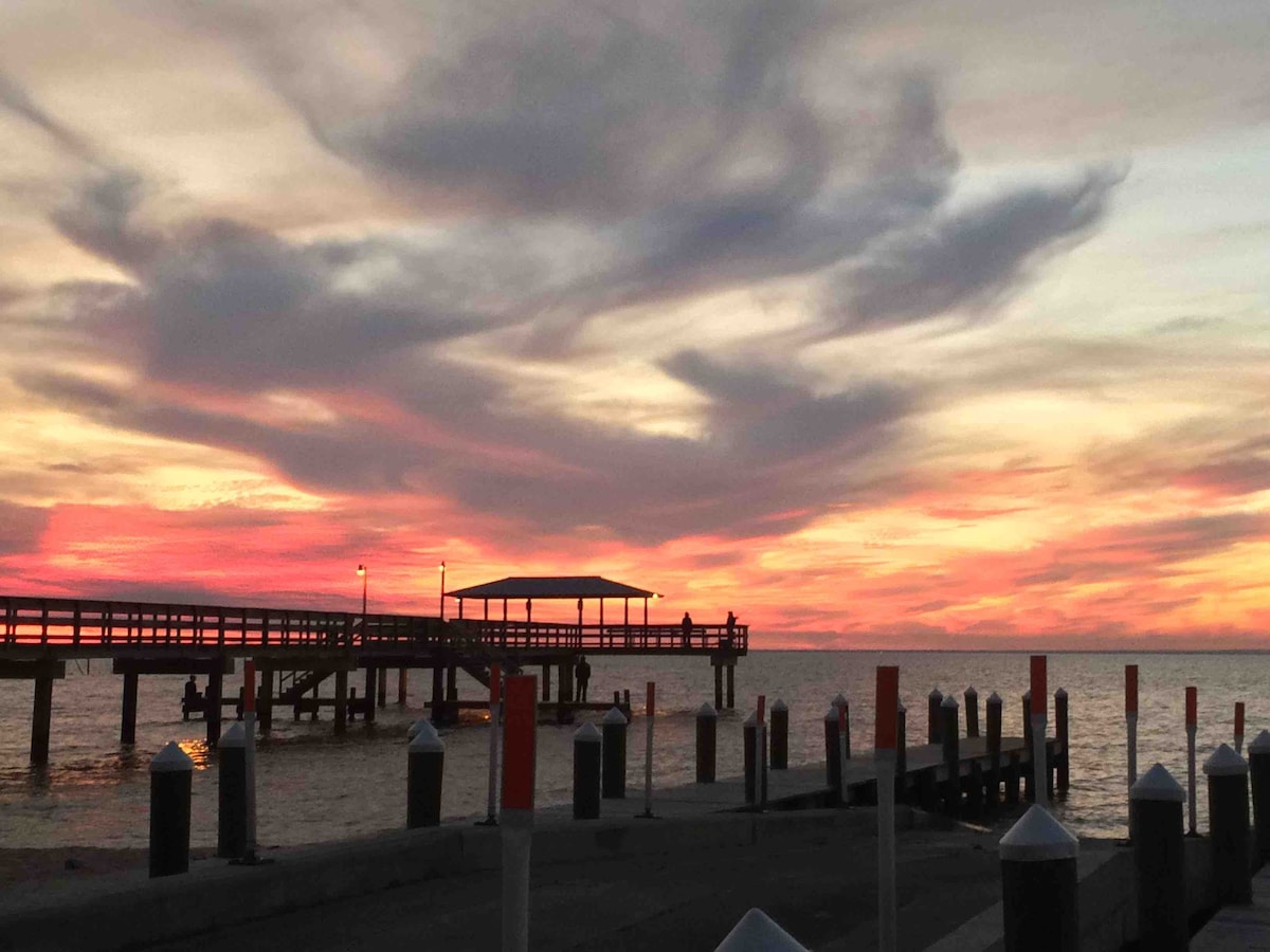 A vibrant sunset paints the sky in shades of orange, pink, and purple above the calm waters. A wooden pier extends into the bay, complemented by reflective pilings and lanterns along the edges, creating a serene coastal scene.