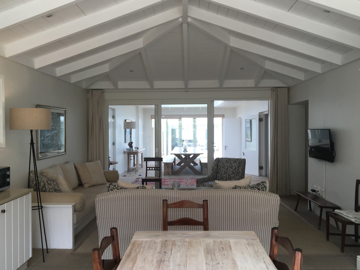 An open-plan living space showcases a light-filled area with high, white-beamed ceilings. Comfortable seating arrangements include a plush sofa and striped chairs. A wooden dining table is positioned in the foreground, while large glass doors lead to a bright, airy room beyond.