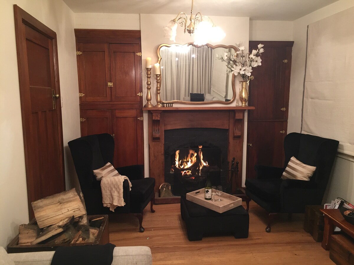 A cozy seating area features two upholstered armchairs positioned near a warm fireplace. A stylish coffee table sits in the center, with a decorative arrangement and a few logs nearby. Vintage cabinetry and a large mirror add character to the welcoming space.