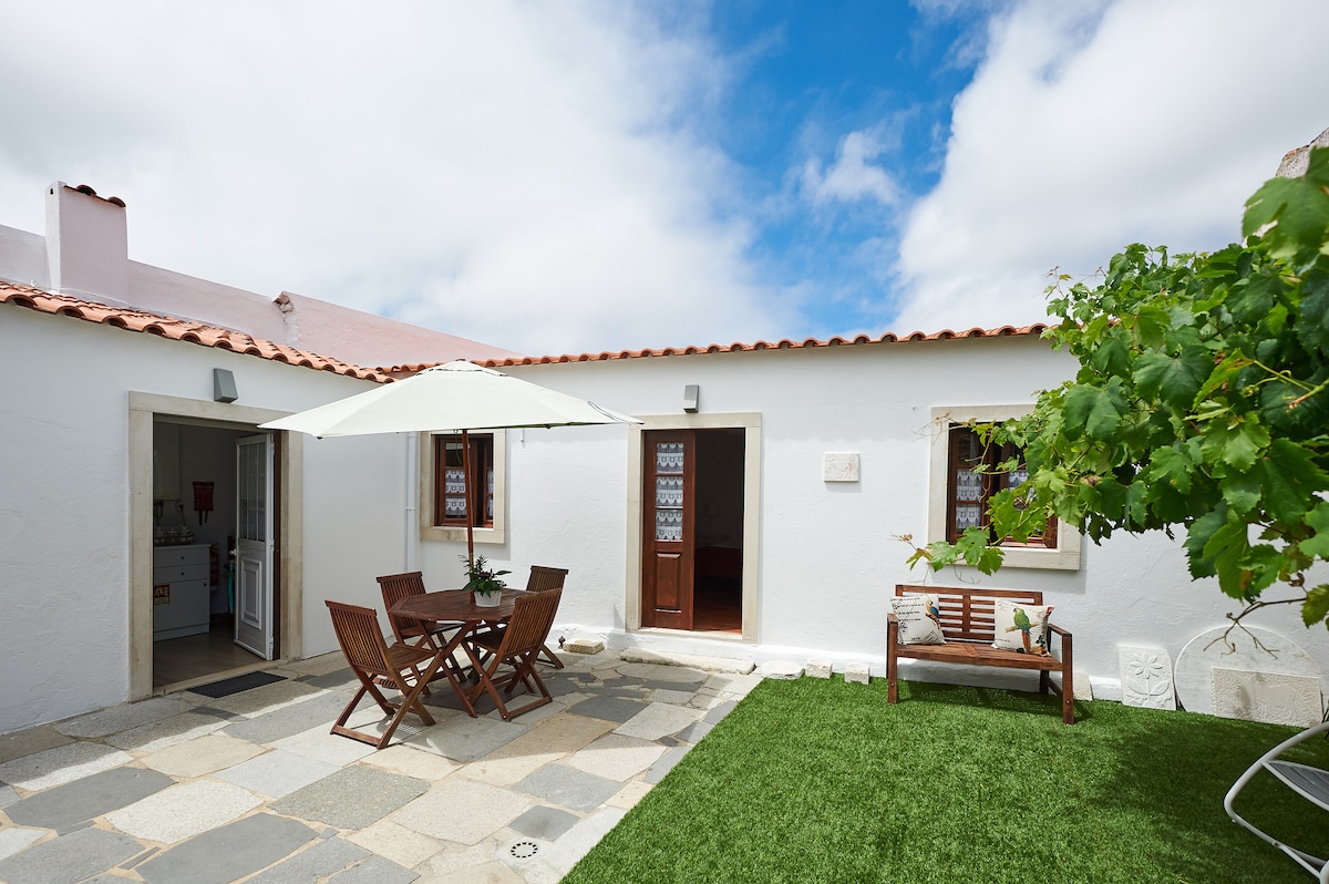 A traditional whitewashed house features a spacious outdoor patio with a wooden table and chairs underneath a large umbrella. Green grass surrounds the seating area, while a small vine-covered trellis adds a natural element. Light clouds drift across a bright blue sky.