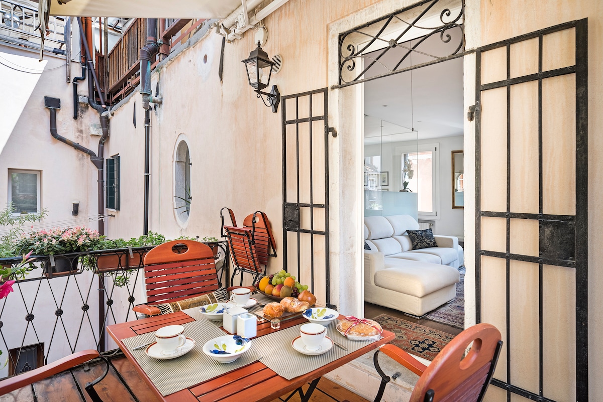 A lovely terrace features a dining table set for breakfast, including plates, cups, and a variety of pastries. Two red chairs enhance the inviting space, with greenery visible in the background. Large doors provide a view into a bright interior living area.